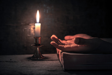 Close up hands praying on bible with light of candle on wooden table at worship room. christian...