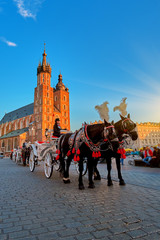 Naklejka na ściany i meble Sunset in Krakow, Polland. Two Horses In Old-fashioned Coach At Old Town Square in beautiful sunset light with St. Mary's Basilica Famous Landmark On Background