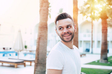 Walking on air. A stunning man with brilliant smile, dark hair and a beard in front of palm trees and white building wearing bright white t-shirt.