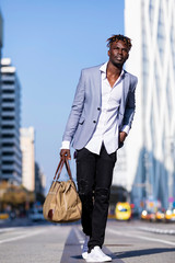 Front view black young man urban casual fashion in the street. Wearing black elegant jacket, jeans, holding and carrying a bag in a sunny day