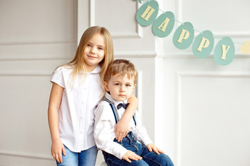 Cute boy in a white shirt is sitting on a chair in a bright room sister hugs his shoulders