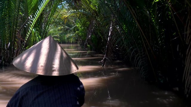Rear view of a local Vietnamese woman wearing a leaf hat and paddling a traditional boat or canoe in the Mekong Delta, Vietnam