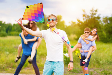 Joyful company of young people in denim clothes are relaxing on green meadow. Smiling man in blue glasses launch colored kite, friends having fun on background. Beautiful nature landscape.