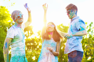 Happy holi party: sunny beautiful woman female teenager and two males men in sunglasses celebrating happy holi festival in spring, summer day outdoor in green park with light leaks and colorful powder