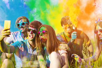 Woman and man soiled with colorful paints, are sitting on grass and making selfie with their...