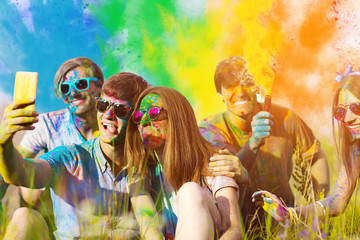 Woman and man soiled with colorful paints, are sitting on grass and making selfie with their friends. Company of young people friends having fun with holi paints and smoke bombs. Holi party concept.