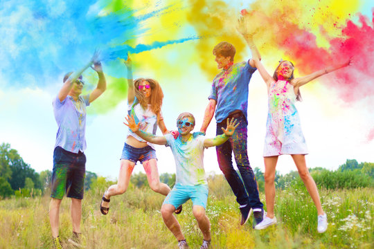 Cheerful and happy friends soiled by bright paints jumping and laugh in colorful smoke on nature. Company of young people having fun with holi paints on spring summer festival. Holi party concept.