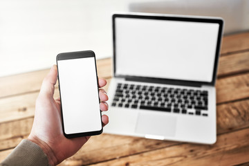 Mobile phone with a blank screen in the male hand palm, in the background a laptop on a wooden table and against a white background