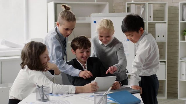 Wide shot of kids surrounded Caucasian boy with tablet in his hands and discussing pictures from internet