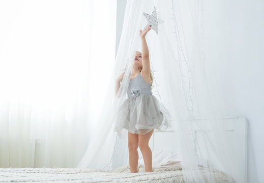 Little toddler girl standing on bed in white bedroom. Cute baby in nice dress tuching a star.