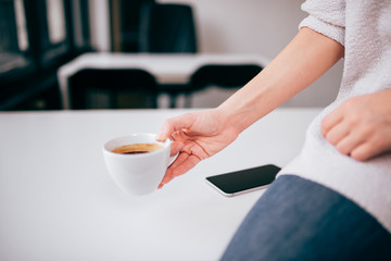 Cropped image of a casual business woman holding a cup of black coffee, sitting on the table with mobile phone.