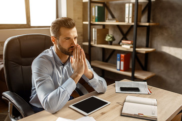 Young handsome thoughtful businessman sit at table in his own office. He hold hands in prayer postition. Thinking. Daylight. Worktime in office.