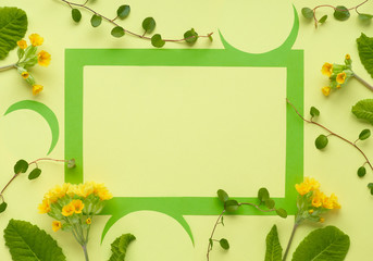 Green paper frame decorated with yellow primrose flowers and leaves, flat lay on yellow paper, text space
