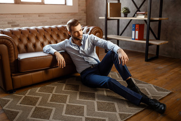 Young handsome businessman sit on floor and pose in his own office. He look straight with confidence. Sexy young man lean to sofa.