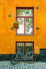 Fototapeta na wymiar Azure vintage bicycle standing in front of vibrant orange building facade wall with big window with red window frame surrounded by several ventilation grills and hanging pot with blooming flowers