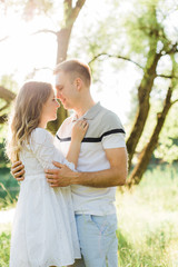 Fototapeta premium Young pretty couple in love standing in park. Handsome cheerful blonde girl in white dress hugging her boyfriend. Man and woman having fun outdoors