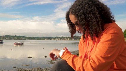 Beautiful mixed race African American girl teenager young woman wearing orange hoodie, sitting on a sea front using her smart watch for social media, communications or texting