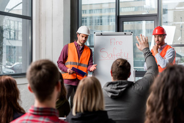 Selective focus of handsome firemen in helmets giving talk on briefing near white board with fire...