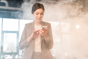 terrified businesswoman looking at smartphone in office with smoke