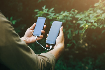 Tourist with smartphone and battery charger is walking in the summer forest. Guy with powerbank is...