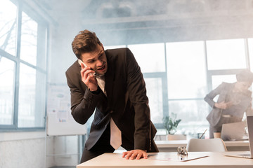 frightened businessman talking on smartphone in office with smoke near female coworker
