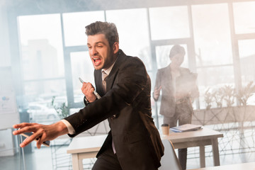 terrified businessman holding smartphone and screaming in office with smoke near female coworker