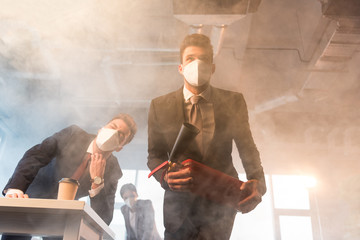  businessman in mask holding extinguisher near coworkers in office with smoke
