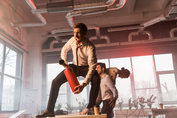 scared businessman yelling while holding extinguisher and standing on desk near coworkers in office...