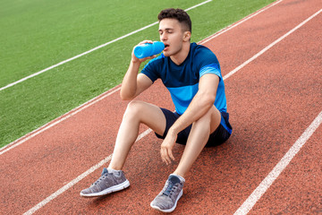 Fototapeta premium Having break. Young man on stadium outside sitting drinking water thirsty closed eyes