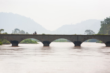 Laotians riding motorcycle crossing ancient bridge over Mekong River.