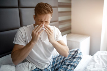 Young man sit on bed early morning. He sneeze into white napkin. Sick and ill young man.
