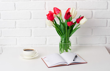 tulip flowers are in a vase on the table, cup of coffee, diary and smartphone, white brick wall as background