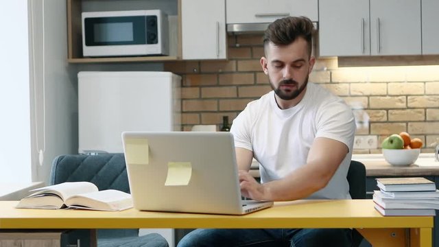 Man learns and writes work on a computer on project using a textbook sitting in the kitchen at home.