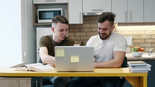 Two guys learn and work on a computer on project using a textbook sitting in the kitchen at home. Talking each other.