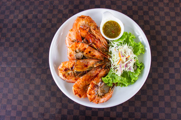 Grilled shrimps with seafood decorated with vegetable and hot spicy dipping sauce in white ceramic dish put on leather floor the grid pattern. Top view