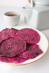 Fototapeta na wymiar Purple dried dragon fruit on white plate with white teapot and teacup in blurred background as snack on white table top cotton fabric texture