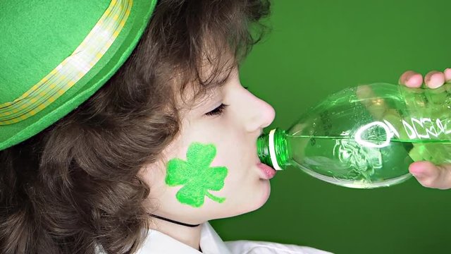 Happy boy with hat and clover image on his cheek drinks water from a bottle isolated on a green background. Profile, Slow Motion.