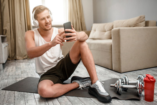 Young well-built man go in for sports in apartment. Happy positive guy sit on carimate on floor and take selfie. Dumbbells and water bottle beside.