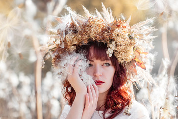 Portrait of a beautiful girl in a white vintage dress and autumn wreath of dried flowers on the head in the midst of airy and fluffy plants. Mysterious fluffy seeds of a Milkweed (Silkweed).