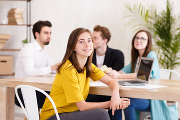 Portrait of successful young woman in casual clothes sitting at an office desk against the background of blurred colleagues - managers of large company