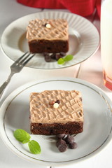 Chocolate bakery style cake brownie  with chocolate cream on top on a white plate and mint leaf and a glass of milk in the background