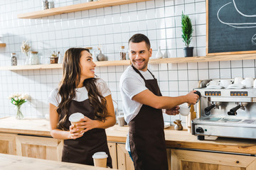handsome barista making coffee and attractive brunette cashier holding paper cup in coffee house