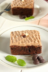 Chocolate bakery style cake brownie  with chocolate cream on top on a white plate and mint leaf and a glass of milk in the background