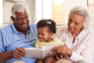 Grandparents Sitting On Sofa At Home Reading Book With Baby Granddaughter