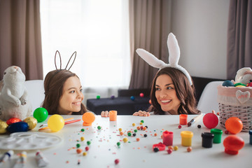 Positive cheerful mother and daughter prepare for Easter. They play and hide behind table in room. Mother and daughter look at each other and smile. They wear bunny ears.