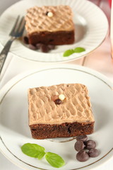 Chocolate bakery style cake brownie  with chocolate cream on top on a white plate and mint leaf and a glass of milk in the background