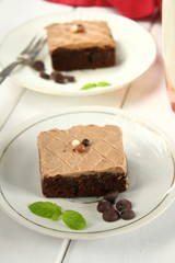 Chocolate bakery style cake brownie  with chocolate cream on top on a white plate and mint leaf and a glass of milk in the background