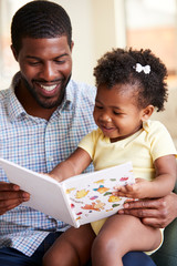 Baby Daughter And Father Sitting On Sofa Reading Book Together