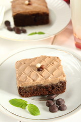 Chocolate bakery style cake brownie  with chocolate cream on top on a white plate and mint leaf and a glass of milk in the background