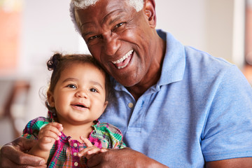 Portrait Of Smiling Grandfather Sitting On Sofa At Home With Baby Granddaughter